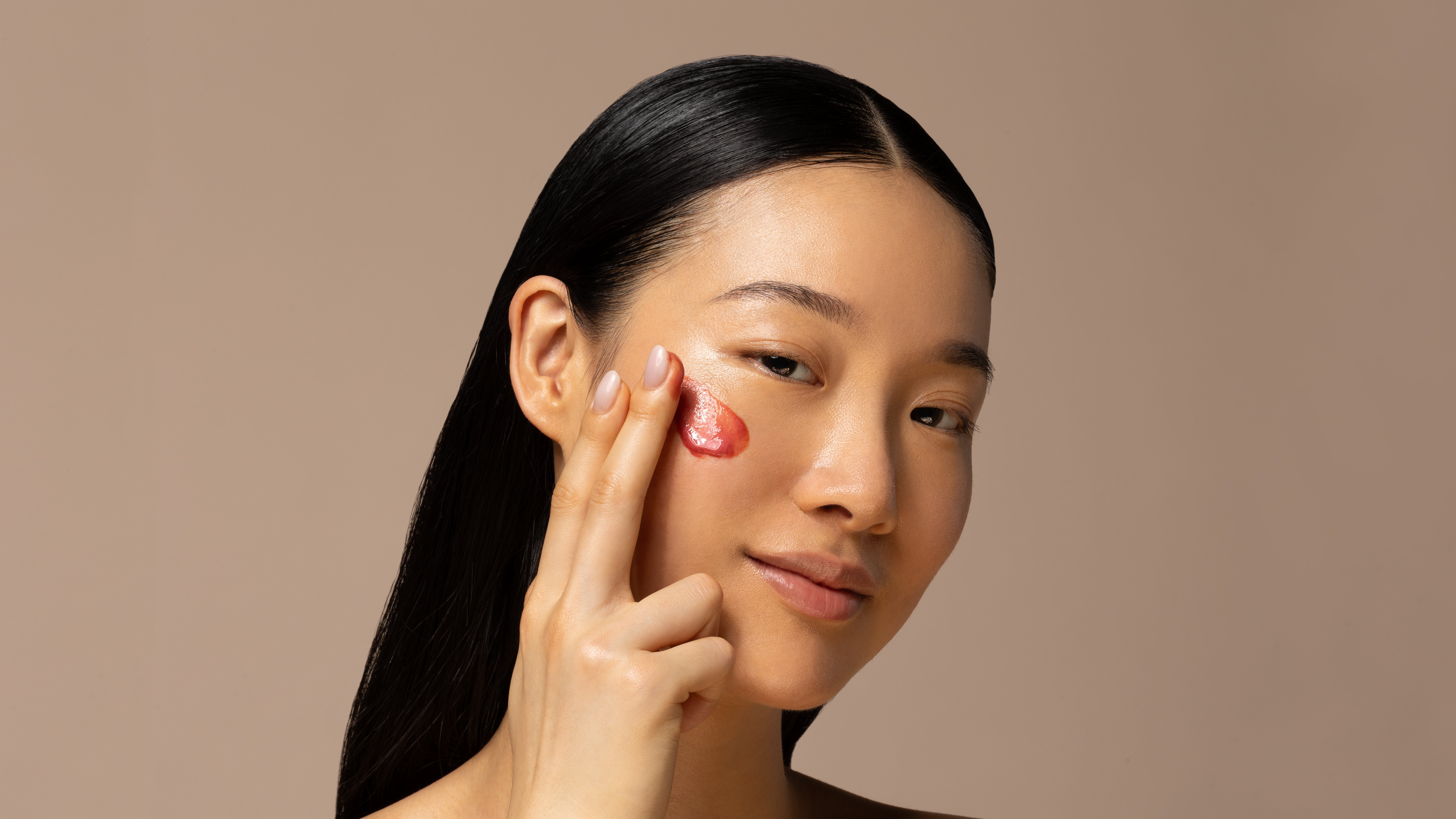 Woman applying a red cream to her cheek against a beige background